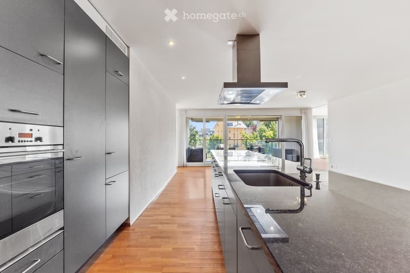Modern kitchen with stainless steel oven, granite countertop, sink, faucet, and grey cabinets. Large windows provide natural light and a view of the outdoor area.
