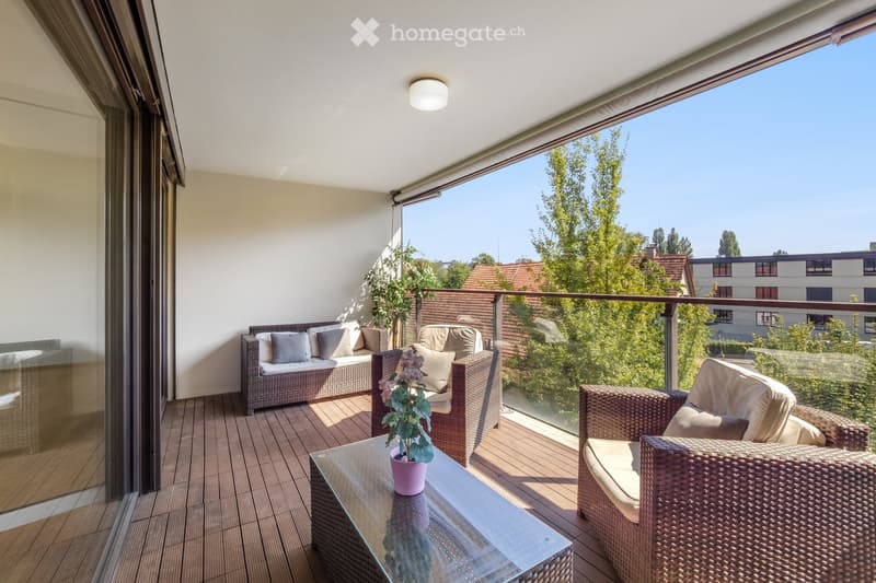 Balcony with wooden decking, wicker furniture including a sofa, chairs, and a glass coffee table with a flower pot, overlooking a green area with trees and buildings.