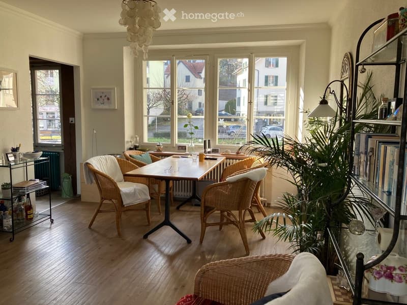 Bright living room with wooden flooring, large windows, a central table with chairs, a floor lamp, and a shelf with books and decorative items.