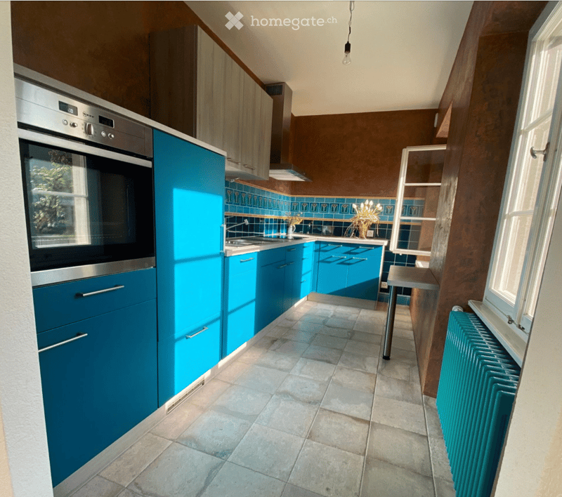 Modern kitchen with blue cabinets, stainless steel oven, tiled backsplash, and a small stool near the window.