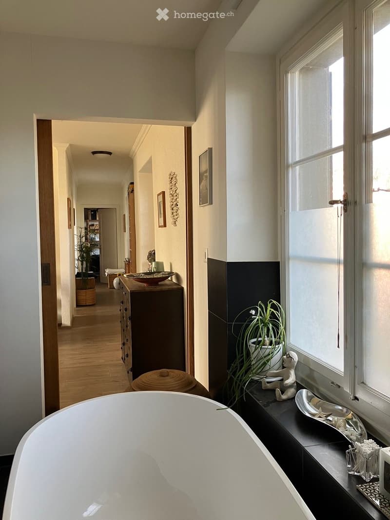 Modern bathroom with a freestanding bathtub, a potted plant on a windowsill, a small stool, a wooden cabinet, and decorative items including a bowl and framed pictures.