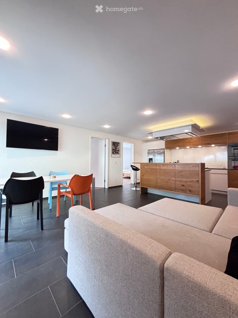 living room with open kitchen, large grey sofa, black and orange plastic chairs, white dining table, black TV on wall, wooden kitchen island, wooden cabinets