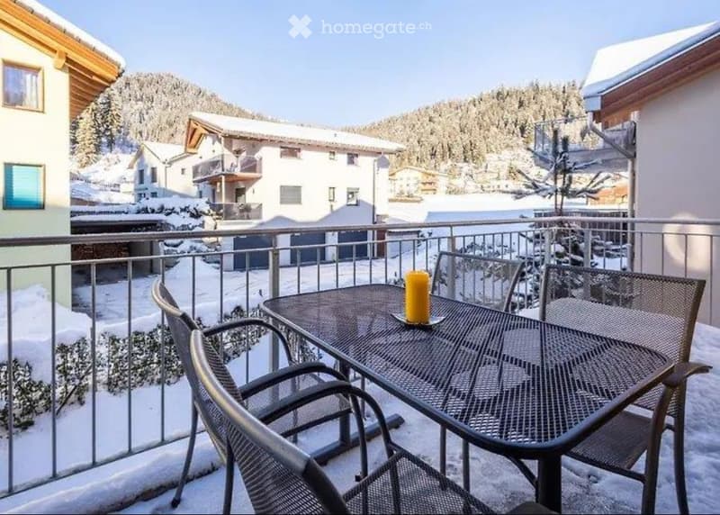 table with chairs on a balcony with a fence, snow on the ground, buildings in the background
