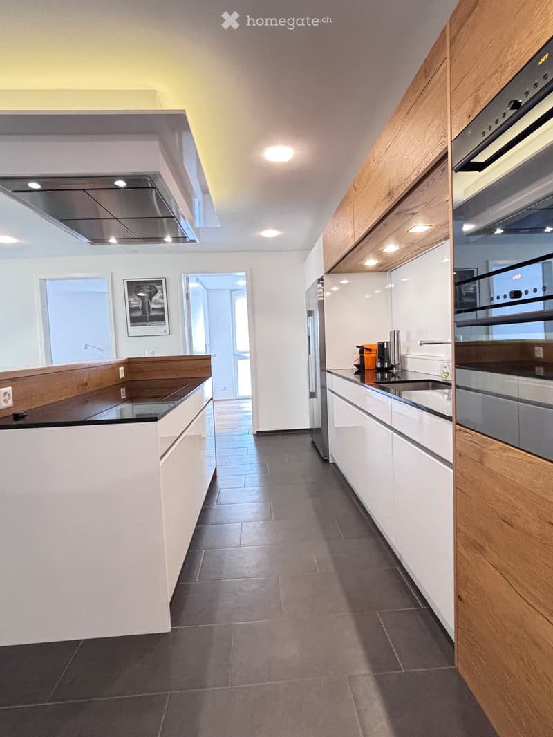 Spacious modern kitchen with wooden cabinets, white countertops, stainless steel appliances, black tiles, large windows, and ceiling lights.