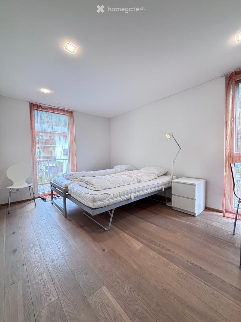 Bedroom with two beds, white walls, wooden floor, bedside table, standing lamp, and curtained window.