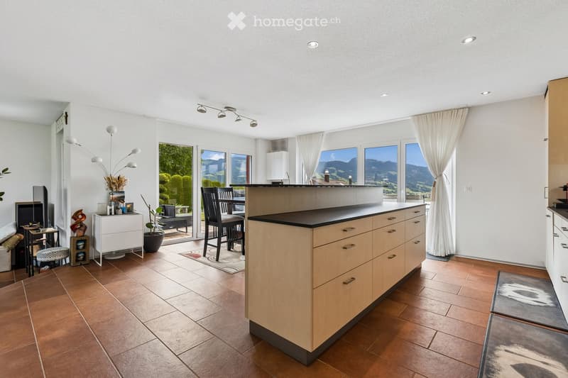 Modern kitchen with light wooden cabinets, a central island, and large windows offering a scenic mountain view. The space includes a dining area with chairs and a table, a potted plant, and decorative items on a side table.