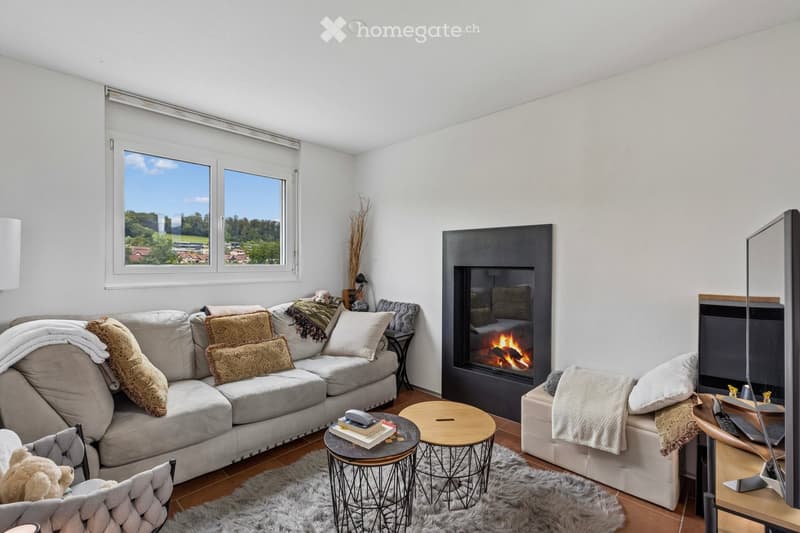 Modern living room with large windows offering a scenic view, a fireplace, a grey sectional sofa, armchairs, coffee tables, and a television.