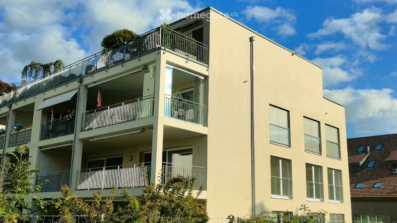 Modern multi-story residential building with balconies, large windows, and a rooftop terrace with greenery.
