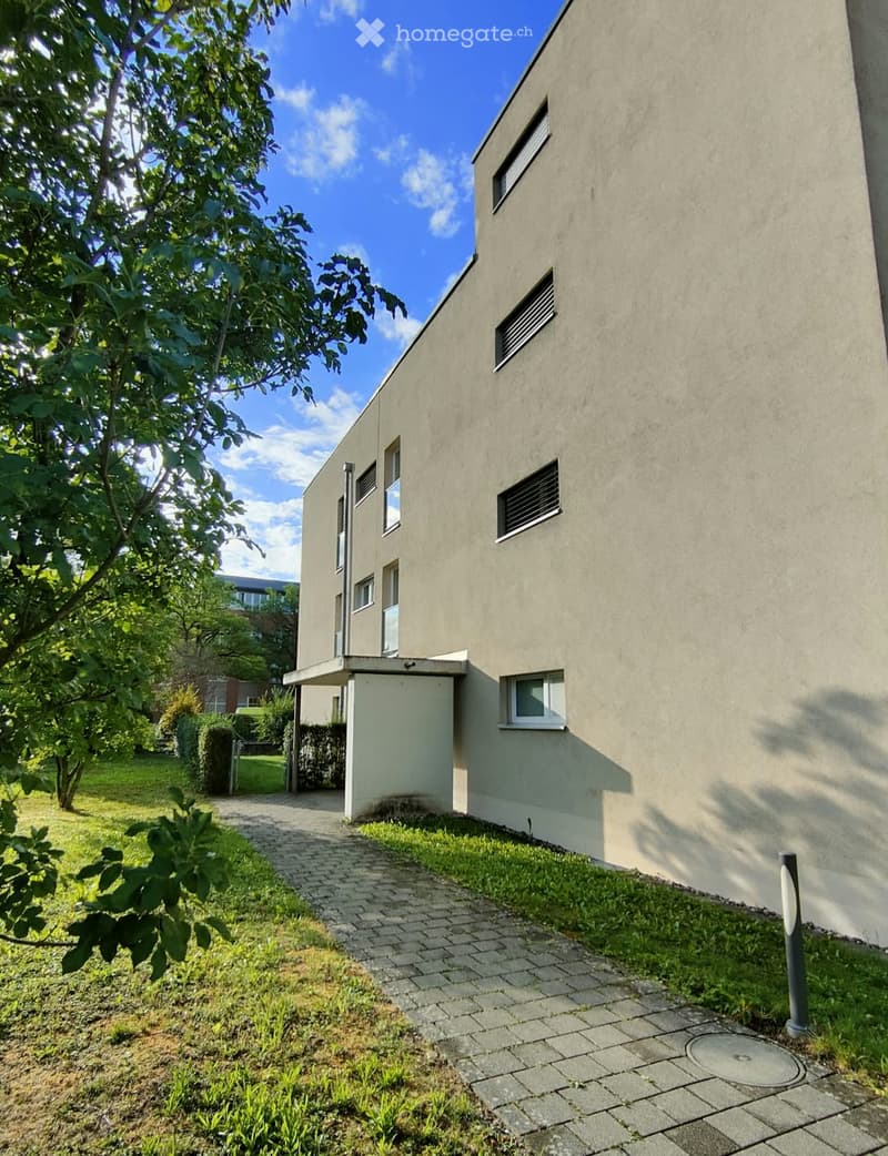 Modern two-story building with a flat roof, concrete facade, and multiple windows. A paved pathway leads to the entrance, surrounded by greenery and trees.