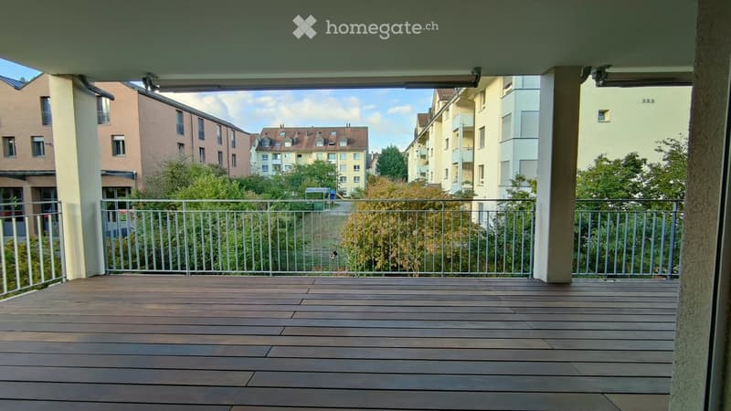 Balcony with wooden decking, metal railing, and view of surrounding buildings and greenery.