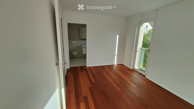 Empty room with wooden flooring, white walls, and a large window with a view of greenery. A small bathroom is visible through an open doorway.