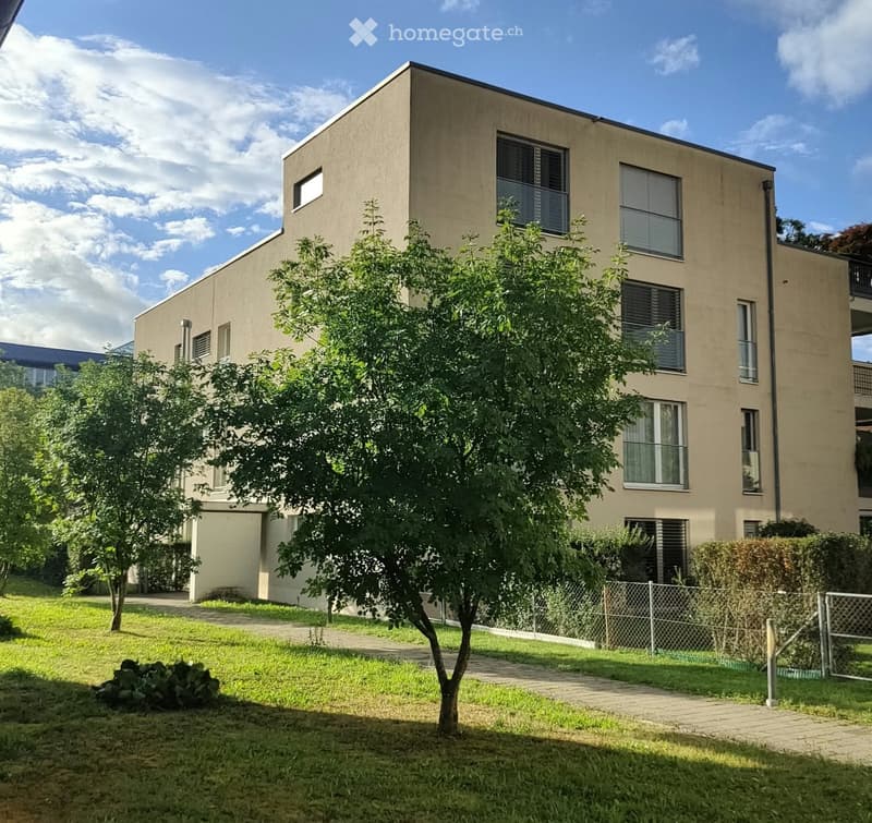 Modern two-story building with beige facade, multiple windows, and a small garden area with a tree and pathway.