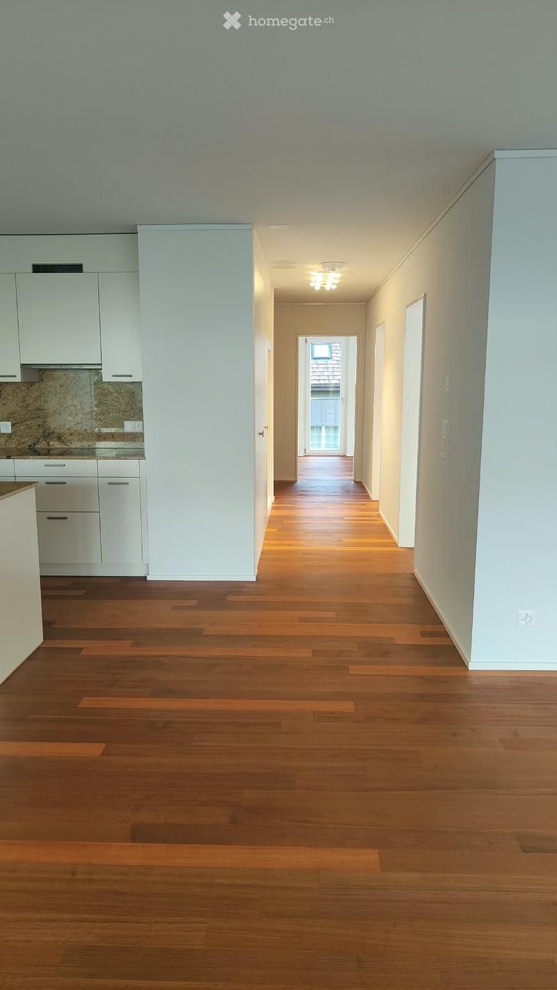 Empty hallway with wooden flooring, white walls, and recessed lighting. The hallway leads to a kitchen area with white cabinets and a granite countertop.
