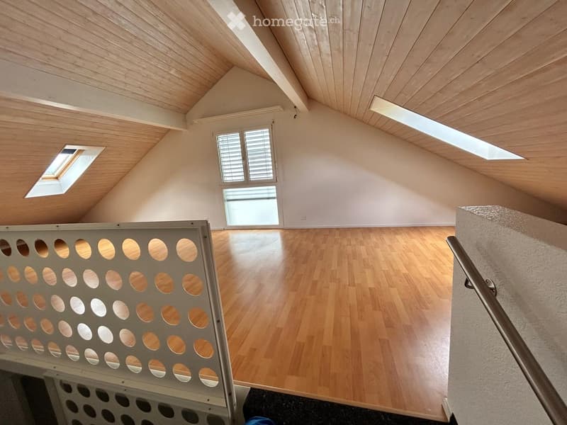 Bright, empty attic space with wooden floors and slanted wooden ceilings. Features large windows and skylights, with a metal staircase leading down.