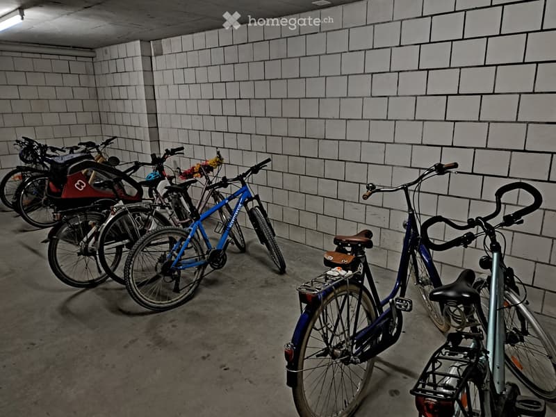 Underground parking area with several bicycles parked against a tiled wall. The space has concrete flooring and exposed brick walls.