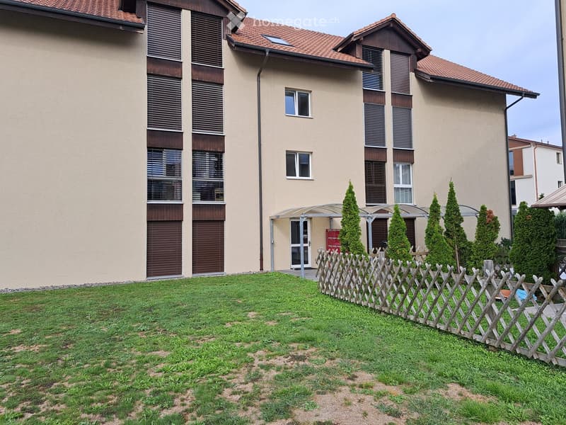 A well-maintained garden area with a wooden fence, neatly trimmed coniferous trees, and a modern house with large windows and a covered entrance.
