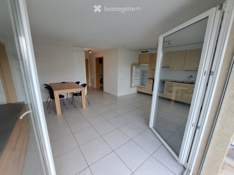 Open-plan living and dining area with tiled flooring, a wooden dining table with four chairs, and a modern kitchen with light wood cabinets and stainless steel appliances.