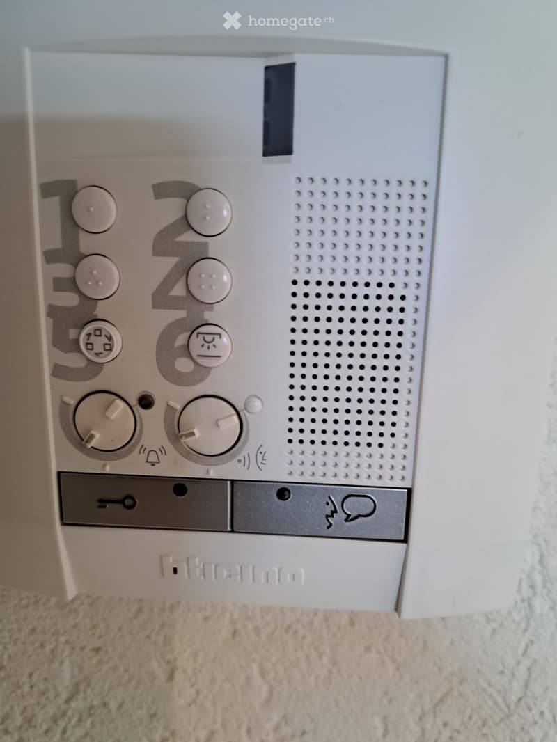 A white thermostat with multiple dials and buttons, mounted on a wall with a textured beige carpet below.