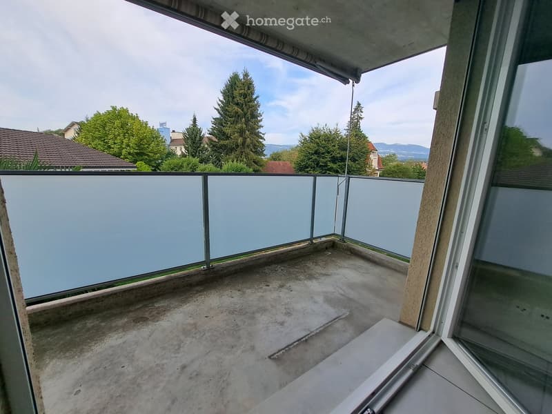 Empty balcony with concrete floor, glass railing, and a view of trees and houses.