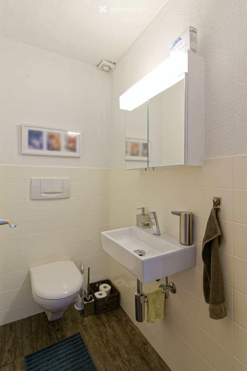 Modern bathroom with white tiles, a toilet, a sink with a modern faucet, a mirror with built-in lighting, a towel, a wicker basket with toilet paper, and a picture frame on the wall.