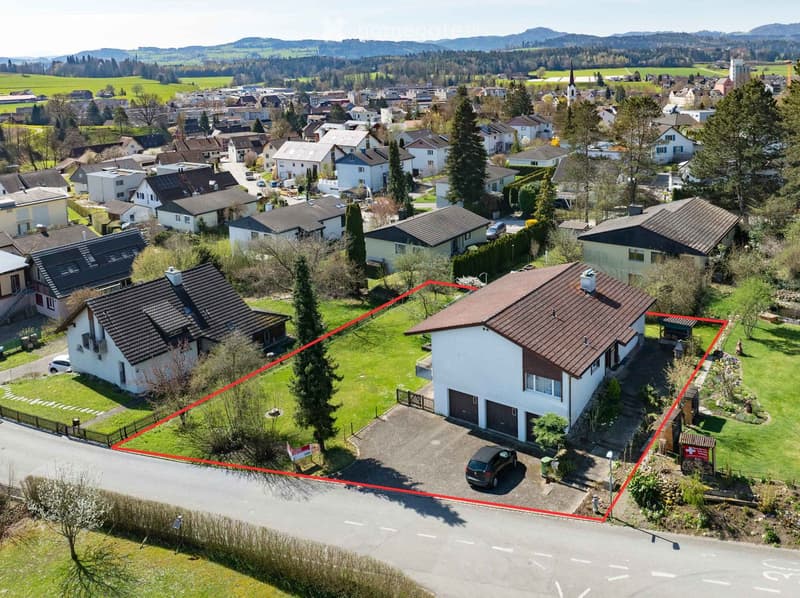 Aerial view of a residential area with multiple houses, green lawns, and a driveway with a parked car. The highlighted house has a garage and a parking area.