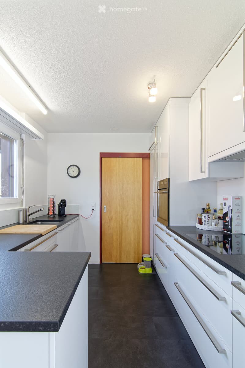 Modern kitchen with white cabinets, dark countertops, and stainless steel appliances. Features include a sink with a faucet, a coffee maker, a cutting board, and a refrigerator. A pet bowl is placed on the floor near the door.