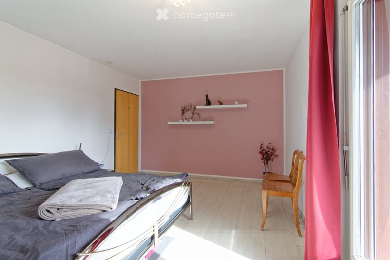 Cozy bedroom with a grey bed, wooden chair, pink accent wall, and decorative shelf with small plants and figurines.