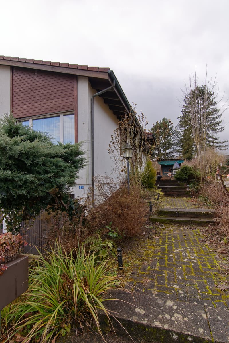 A garden area with a stone pathway leading to a house entrance, surrounded by various plants and shrubs, with a small outdoor seating area visible in the background.