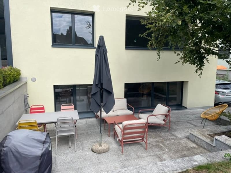 A modern patio area with a table, chairs, and a large umbrella, adjacent to a house with large windows and a sliding glass door.