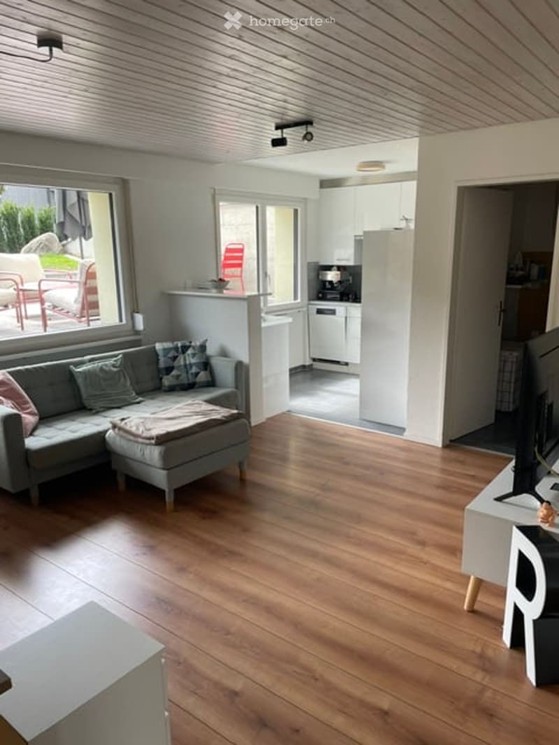 Bright living room with wooden flooring, large windows, grey sectional sofa, ottoman, coffee table, and a small dining area with a table and chairs. A modern kitchen is visible in the background with white cabinets and stainless steel appliances.