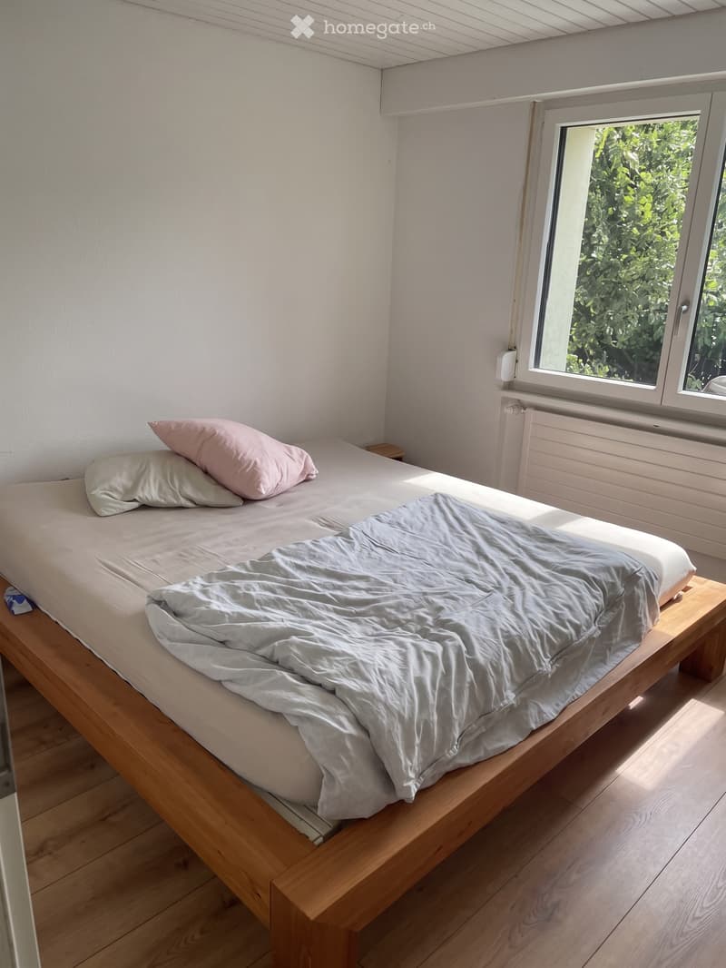 Cozy bedroom with a wooden bed frame, light grey bedding, and a pink pillow, situated near a window with a view of greenery.