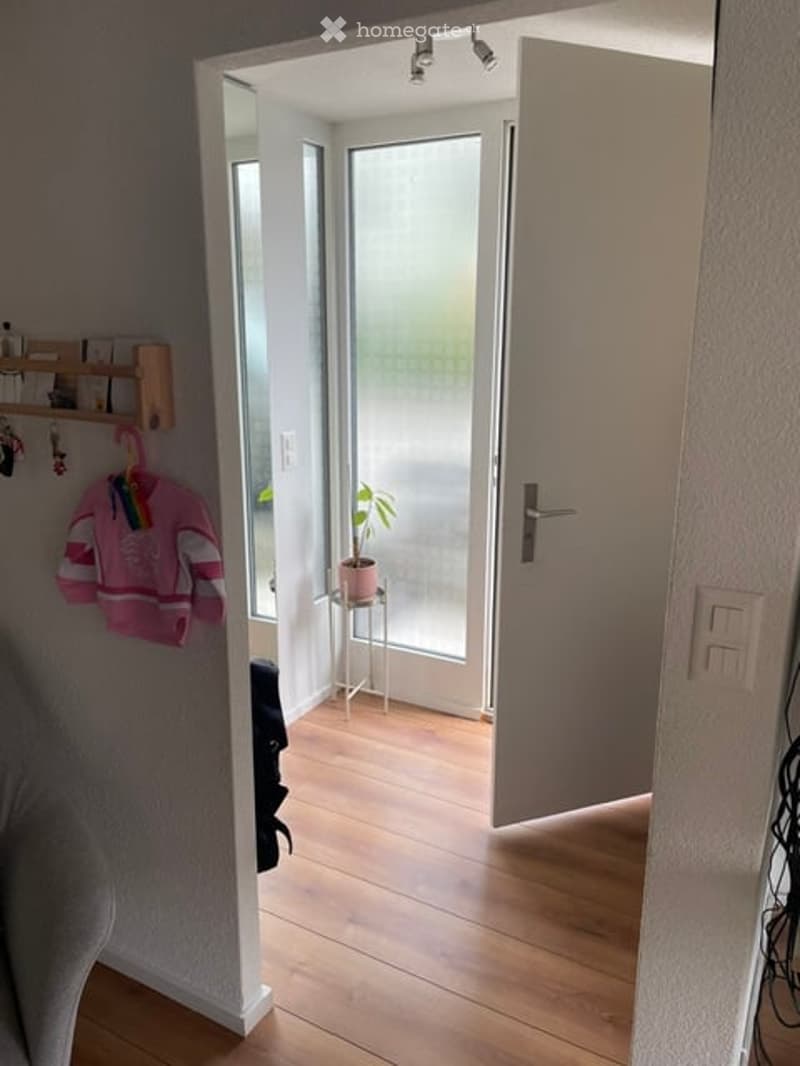 A narrow hallway with wooden flooring, white walls, and a partially open door leading to a bright room with a small plant on a table.