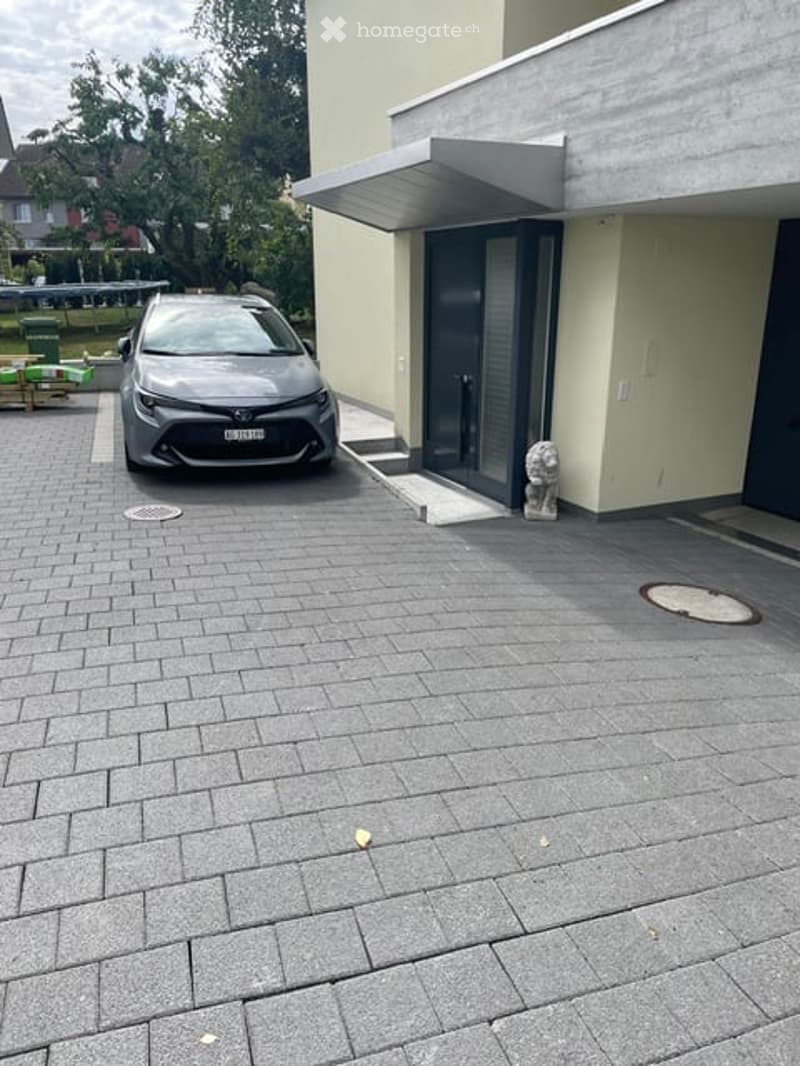 Driveway with a parked car, modern house entrance, and a small garden area with outdoor furniture.