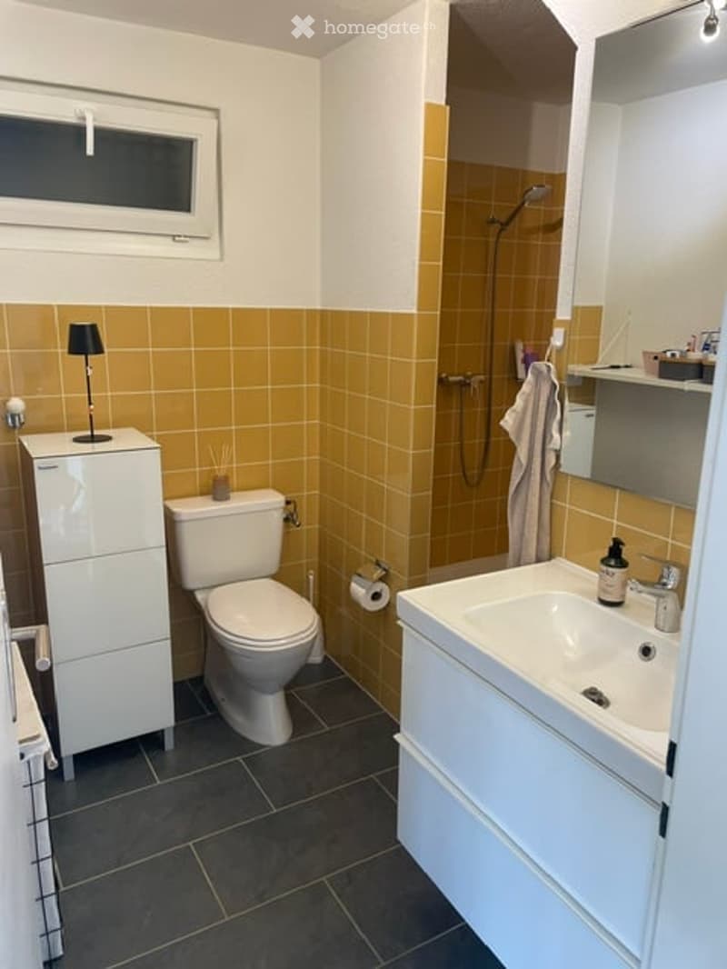 Modern bathroom with yellow and white tiles, a toilet, a sink with a mirror, a shower, and a white cabinet with a lamp on top.