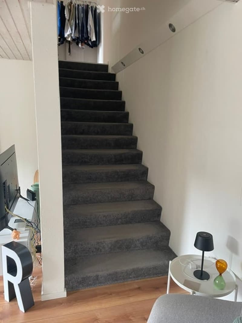 Modern hallway with a carpeted staircase, wooden flooring, and a small round table with a lamp and decorative items on the right side.