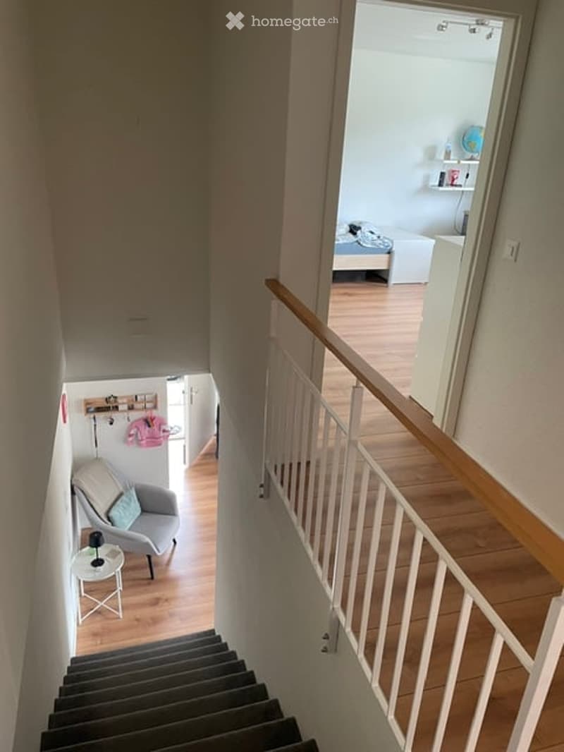 A modern hallway with wooden flooring, white walls, and a staircase leading to an upper level. The hallway features a grey armchair with a blue pillow, a small white side table with a lamp, and a coat rack with a pink jacket. The upper level shows a glimp
