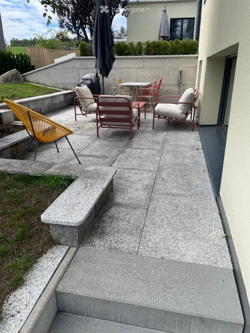 Modern outdoor patio with concrete flooring, several chairs and a table, umbrella, and a raised bench.