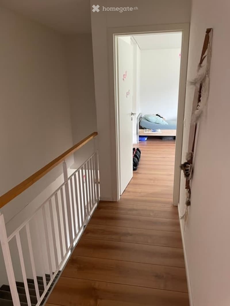 A narrow hallway with wooden flooring and white walls, leading to an open doorway that reveals a small bedroom with a bed and a nightstand.