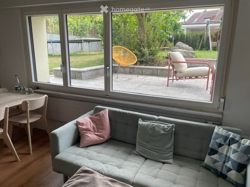 Bright living room with large windows overlooking a garden, featuring a grey sofa with colorful pillows, a small dining table with chairs, and outdoor seating visible through the windows.