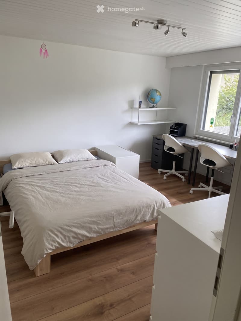 Modern, compact bedroom with a bed, desk, chair, and globe on a shelf. The room features light wooden flooring and white walls.