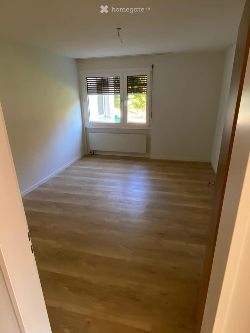 Empty room with light wooden flooring, white walls, and a window with blinds. A radiator is visible below the window.