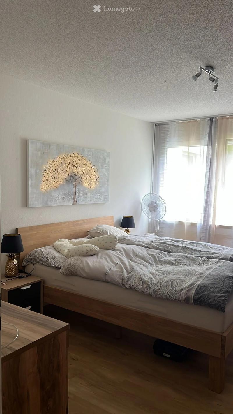 Cozy bedroom with a wooden bed, nightstands, lamps, and a fan near the window. A large abstract painting adorns the wall.
