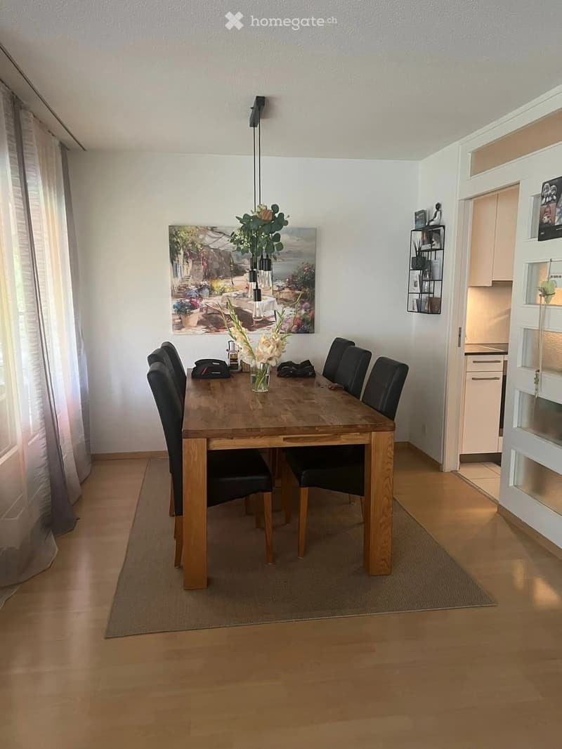 Dining room with wooden dining table, four black chairs, vase with flowers, painting on wall, and a shelf with items visible in the background.
