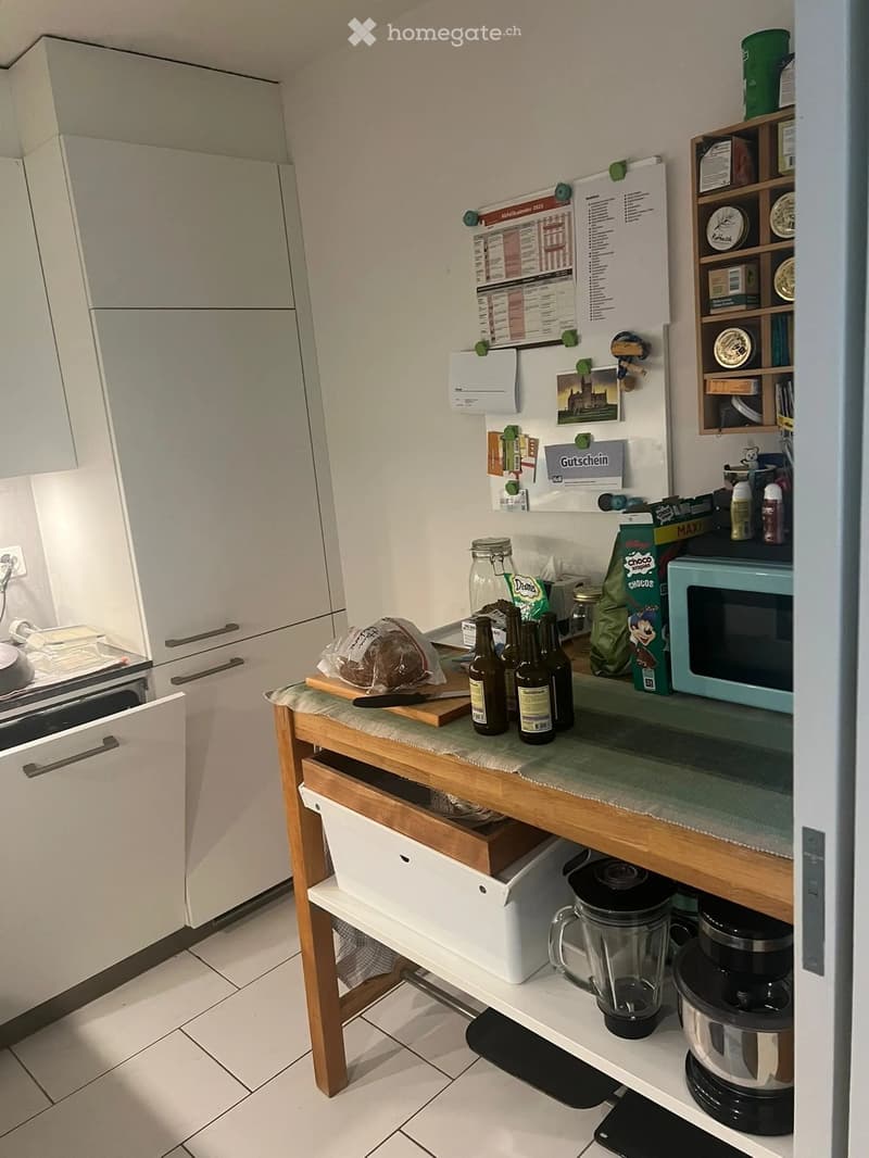 Modern kitchen with white cabinets, a wooden island, and various appliances including a refrigerator, microwave, blender, and coffee maker. The island has a countertop with bottles, a cutting board, and a loaf of bread.