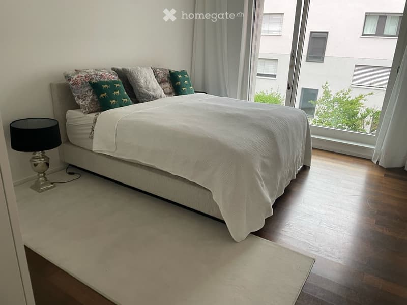 Modern bedroom with a large bed, decorative pillows, a bedside lamp, and large windows with a view of greenery outside.