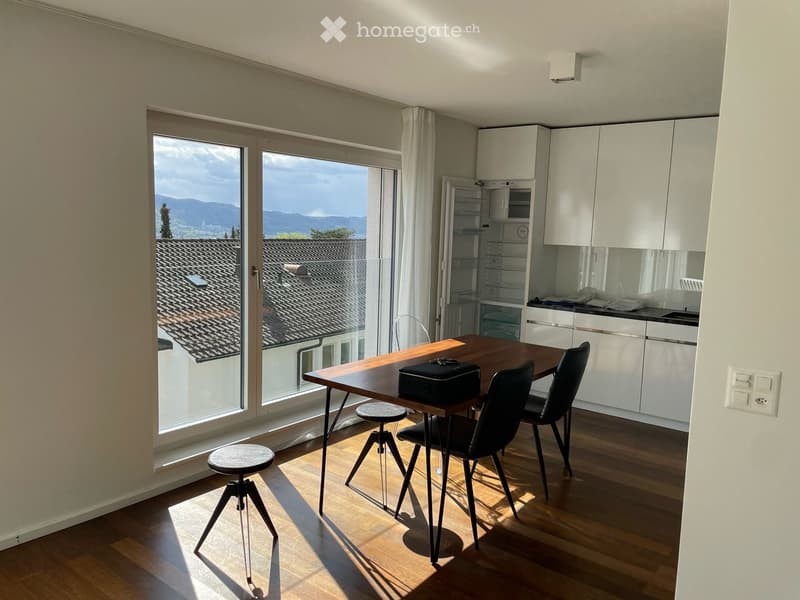 Bright dining area with wooden table, chairs, and a stool, adjacent to a modern kitchen with white cabinets and a refrigerator, large windows offering a scenic view of rooftops and mountains.