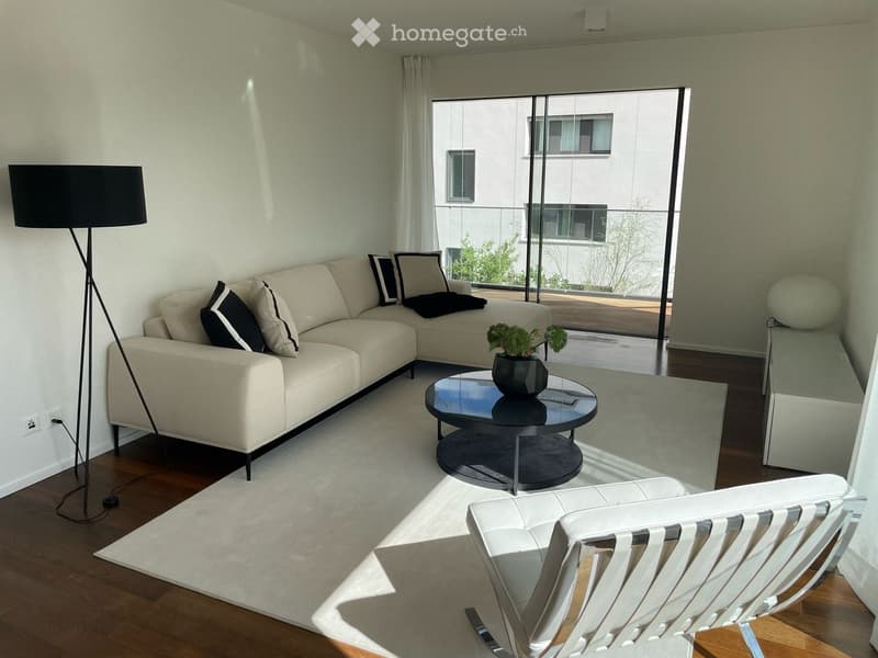 Modern living room with a white sectional sofa, black and white throw pillows, a round black coffee table with a potted plant, a white modern chair, and a tall floor lamp. Large windows and a balcony are visible.