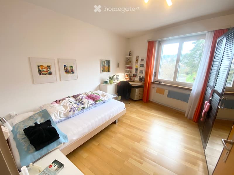 Bright bedroom with wooden flooring, a single bed with colorful bedding, a desk with a chair, a small plant, framed pictures on the wall, and a large window with orange curtains.