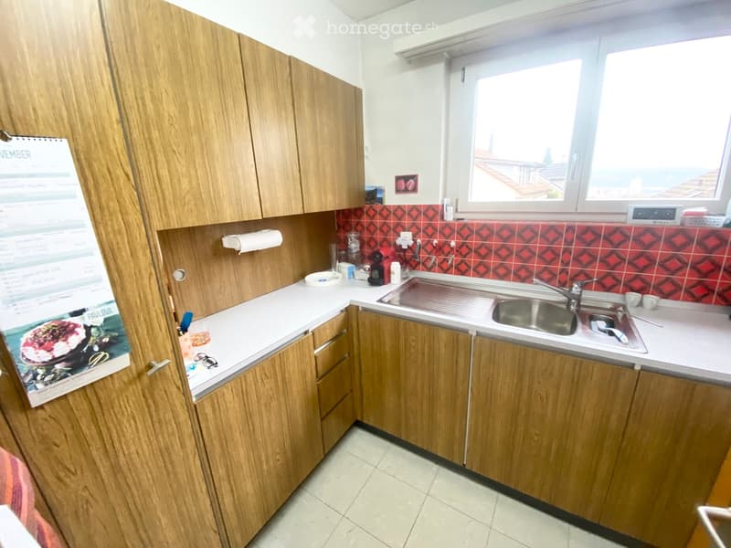 A compact kitchen with wooden cabinets, a white countertop, and a stainless steel sink. The backsplash features a red and black tile design. A window offers a view of the outside.