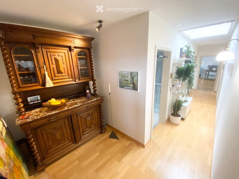 Bright hallway with wooden flooring, a large wooden cabinet with glass doors, a fruit bowl, and potted plants. The hallway leads to another room with visible furniture.